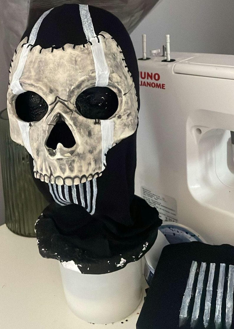 May include: A skull mask with a black hood. The skull is painted in a bone-like colour with black eye sockets and a large nose opening. White stripes accent the skull and the hood. The mask is likely for Halloween or cosplay.