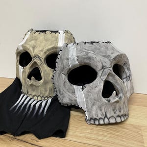 May include: Two skull masks with stitched seams and painted details. One mask is tan with a white stripe, the other is grey with a white stripe. A black fabric with white painted lines is beneath the masks.