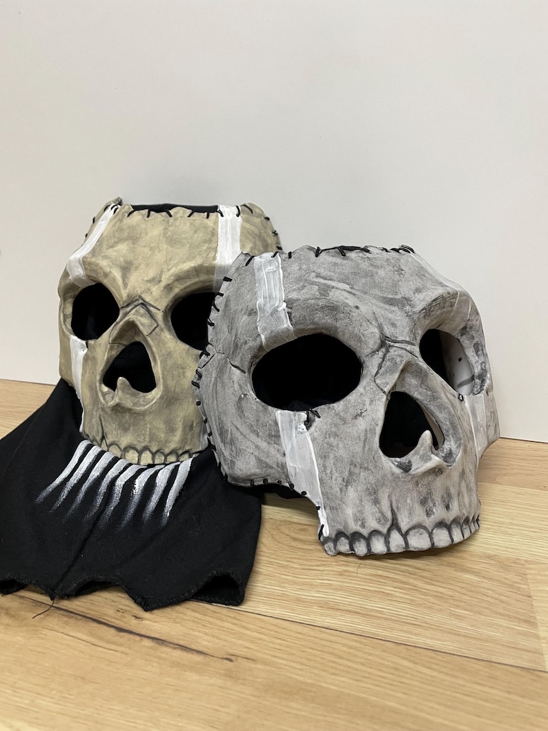 May include: Two skull masks with stitched seams and painted details. One mask is tan with a white stripe, the other is grey with a white stripe. A black fabric with white painted lines is beneath the masks.