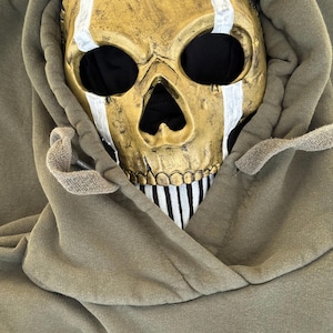 May include: A gold skull mask with white stripes and black eye sockets sits within the hood of an olive green hoodie. The mask has a stitched design and a black and white striped pattern on the lower jaw. The hoodie has the letters "MP" on the front.