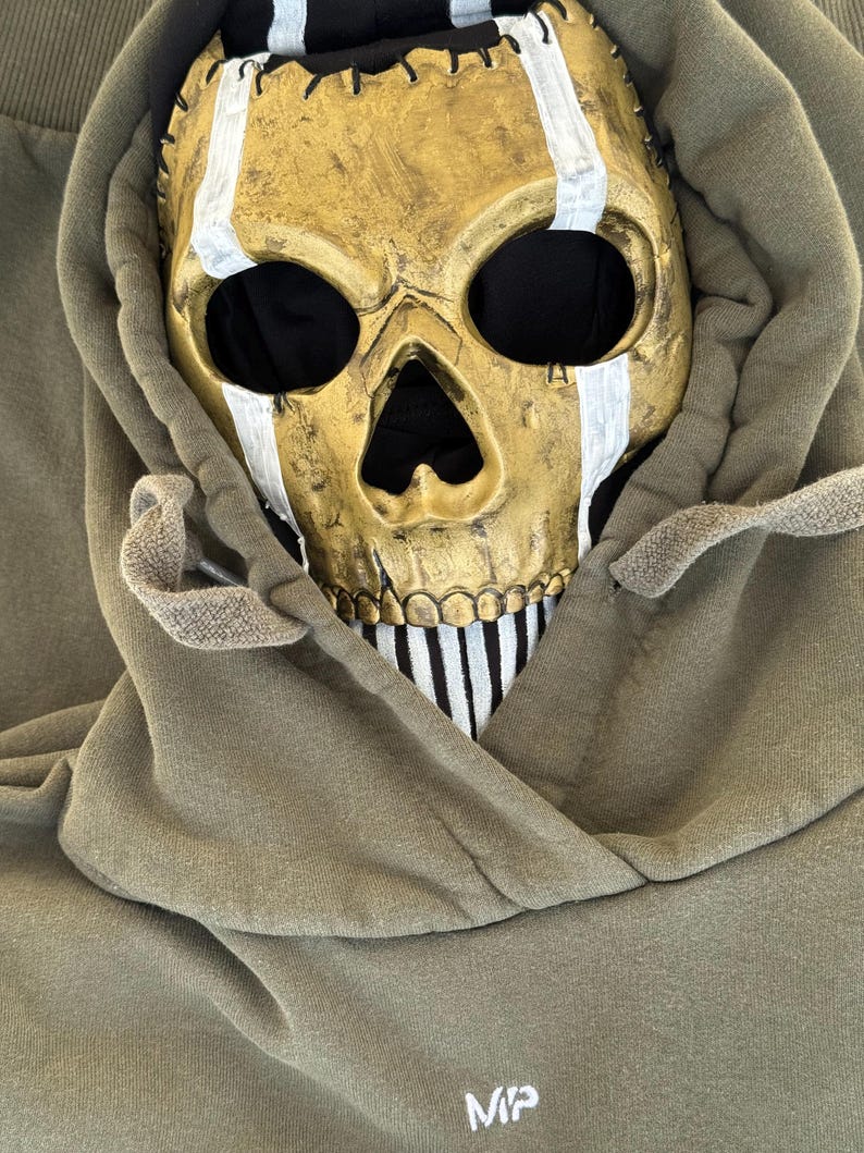 May include: A gold skull mask with white stripes and black eye sockets sits within the hood of an olive green hoodie. The mask has a stitched design and a black and white striped pattern on the lower jaw. The hoodie has the letters "MP" on the front.
