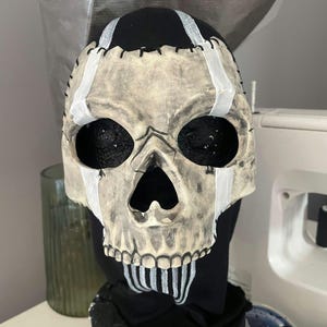 May include: A skull mask with a distressed, aged appearance. The mask features black eye sockets and a detailed skeletal design. White paint accents the forehead and jawline, with black stitching details. The mask is set against a black fabric background.