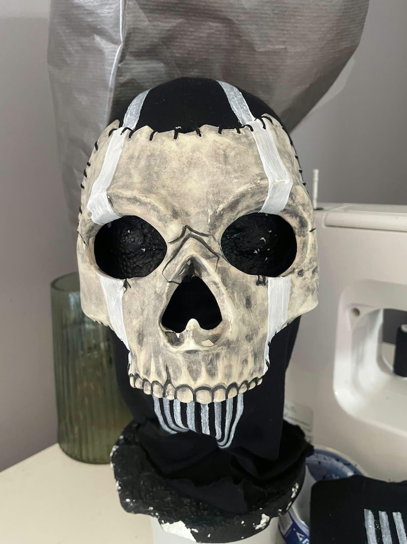 May include: A skull mask with a distressed, aged appearance. The mask features black eye sockets and a detailed skeletal design. White paint accents the forehead and jawline, with black stitching details. The mask is set against a black fabric background.