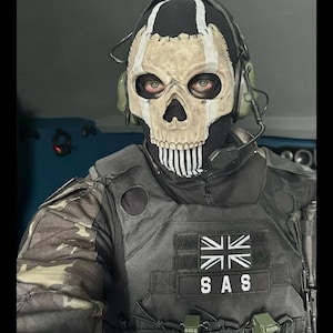 May include: A person wearing a skull mask with a black hood and tactical vest. The mask has a white skull design with black accents. The vest has a Union Jack patch and the letters "SAS". Two green grenades are attached to the vest.