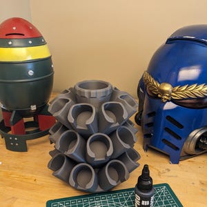 May include: A gray, 3D-printed paint holder with multiple cylindrical slots, next to a blue helmet with gold accents and a red, yellow, and green bomb-shaped object. A bottle of black paint sits on a green cutting mat.