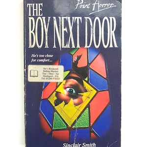 May include: A book cover with a dark blue background and the title "The Boy Next Door" in white letters. The subtitle is "He's too close for comfort..." and the author is "Sinclair Smith". The cover features a stained glass window with a single eye peering through a broken pane.