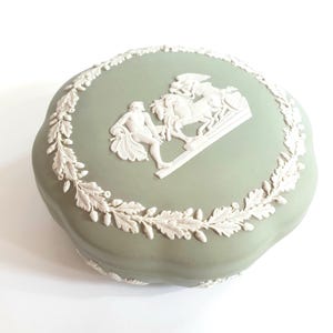 Può includere: Una scatola portaoggetti in ceramica verde e bianca con un design in rilievo di una scena classica. La scatola è decorata con un bordo bianco di foglie e ghiande.