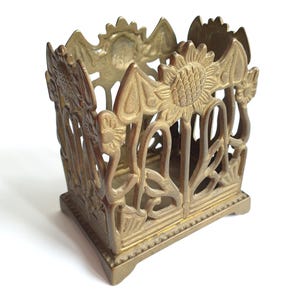 May include: A decorative brass-colored metal container with a square base and openwork sides. The sides feature a floral design with sunflowers and stylized leaves. The container is likely for holding small items or as a decorative piece.