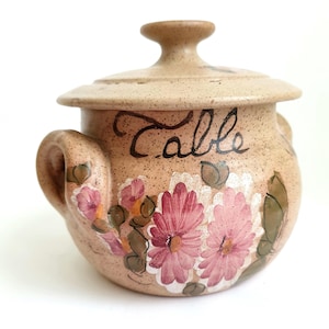 May include: A tan ceramic container with a lid, handles, and floral design. The container has the word "Table" written on it in black script. Pink and white flowers are painted on the front.