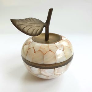 Puede incluir: Caja de baratijas decorativa con forma de manzana. El cuerpo está cubierto de un mosaico de azulejos iridiscentes de color crema con una banda de latón en el medio. Un tallo y una hoja de latón coronan la manzana, añadiendo elegancia. Un objeto decorativo único.