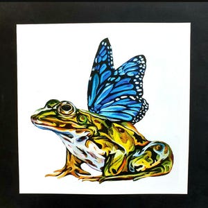 Puede incluir: Una rana verde con un ala de mariposa azul en la espalda. La rana está sentada sobre un fondo blanco.