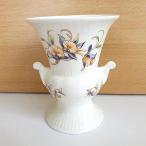 May include: White ceramic vase with a flared top and two small handles. The vase features a floral design in shades of blue, yellow, and orange. The base has a ribbed texture. A decorative piece for the home.