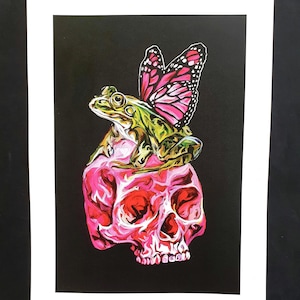Puede incluir: Una rana verde con una mariposa rosa en la espalda se sienta encima de una calavera rosa sobre un fondo negro.