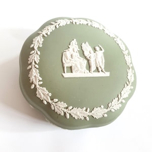 Peut inclure: Une boîte à bijoux en céramique verte avec un motif blanc en relief de trois personnages. La boîte est de forme ovale avec un bord festonné et une bordure florale blanche.