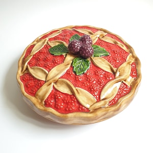 May include: A round ceramic pie dish with a lid featuring a sculpted red berry filling, a tan lattice crust, and three dark purple blackberries with green leaves on top. The surface has a glossy finish with realistic textures on the fruit.