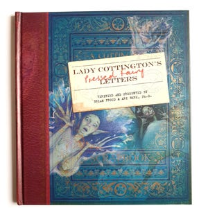 May include: A book titled "Lady Cottington's Pressed Fairy Letters" with a blue cover and gold detailing. The spine is red. A cream-colored label is attached, displaying the title in red and black. The cover features illustrations of fairies, creating a whimsical aesthetic.
