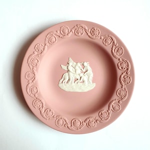 Può includere: Un piatto decorativo rosa con un design a bassorilievo bianco. Il piatto presenta una scena centrale di figure con un cavallo alato, circondata da un bordo dettagliato. Il piatto è di colore rosa chiaro.