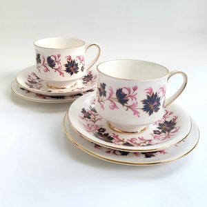 Könnte beinhalten: Zwei weiße Teetassen und Untertassen mit einem floralen Muster in Rosa-, Blau- und Goldtönen. Die Tassen und Untertassen haben einen goldenen Rand.