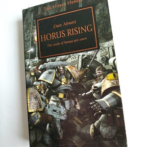 Puede incluir: Un libro de bolsillo titulado "Horus Rising" de Dan Abnett. La portada presenta una ilustraci&oacute;n oscura y llena de acci&oacute;n de figuras blindadas en una escena de batalla. El libro es parte de la serie "The Horus Heresy".