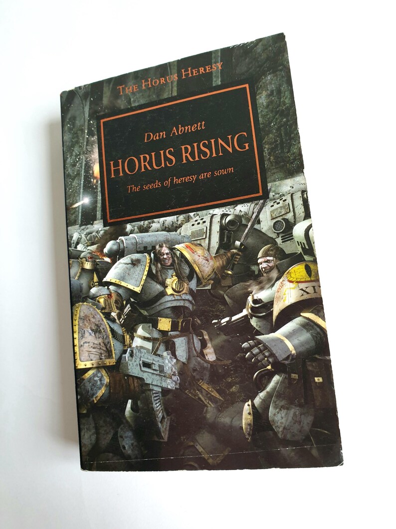 Puede incluir: Un libro de bolsillo titulado "Horus Rising" de Dan Abnett. La portada presenta una ilustraci&oacute;n oscura y llena de acci&oacute;n de figuras blindadas en una escena de batalla. El libro es parte de la serie "The Horus Heresy".