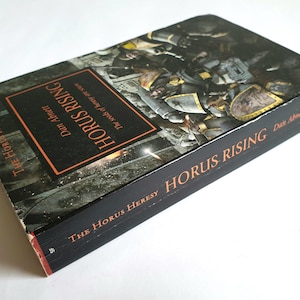 Puede incluir: Un libro de tapa dura titulado "Horus Rising" de Dan Abnett. El libro tiene una cubierta negra con texto naranja y una ilustraci&oacute;n gr&aacute;fica. Forma parte de la serie "The Horus Heresy".