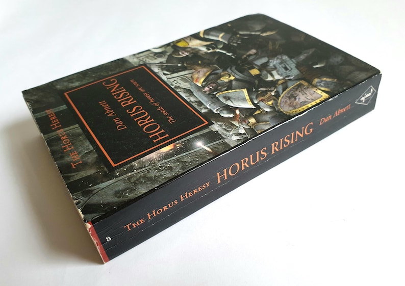 Puede incluir: Un libro de tapa dura titulado "Horus Rising" de Dan Abnett. El libro tiene una cubierta negra con texto naranja y una ilustraci&oacute;n gr&aacute;fica. Forma parte de la serie "The Horus Heresy".