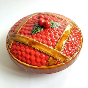 Puede incluir: Un plato redondo de cerámica con tapa en forma de tarta de cerezas. La tapa presenta una superficie texturizada de bayas rojas con un patrón de corteza de rejilla marrón brillante y un grupo central de tres cerezas rojas con dos hojas.