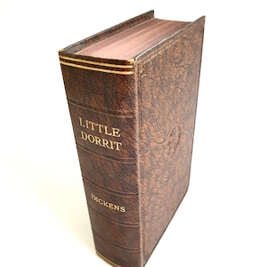 May include: A vintage copy of "Little Dorrit" by Dickens. The book features a brown, textured cover with gold lettering and detailing. The pages are aged, and the spine is visible.