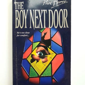 May include: A paperback book titled "The Boy Next Door" with the subtitle "He's too close for comfort..."  The cover features a stained glass window with a single eye peering through a broken pane. The book is part of the Point Horror series and is written by Sinclair Smith.