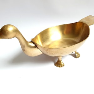 May include: A polished brass bowl shaped like a duck, featuring a long neck, an open beak, a wide oval body, a flat tail, and two webbed feet. The metallic surface has a warm golden hue and a smooth finish, creating a functional vessel.