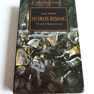 Puede incluir: Un libro de bolsillo titulado "Horus Rising" de Dan Abnett. La portada presenta una escena de batalla con figuras blindadas en tonos grises, dorados y amarillos. El libro forma parte de la serie "The Horus Heresy", con el lema "The seeds of heresy are sown."