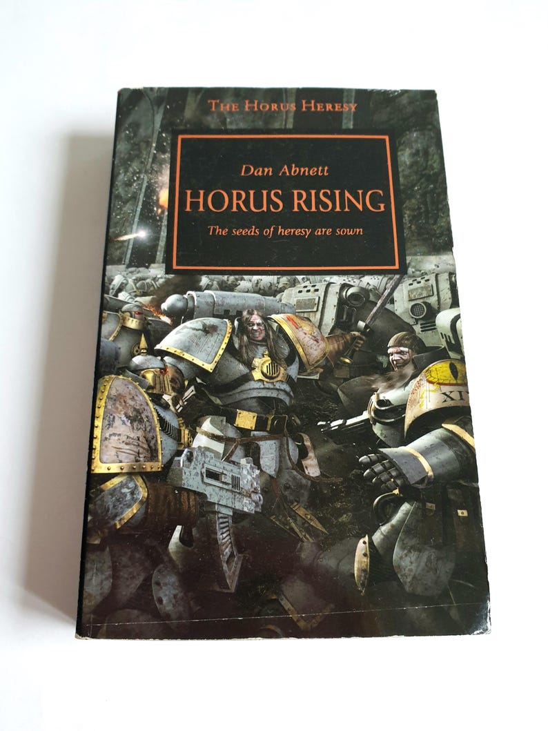 Puede incluir: Un libro de bolsillo titulado "Horus Rising" de Dan Abnett. La portada presenta una escena de batalla con figuras blindadas en tonos grises, dorados y amarillos. El libro forma parte de la serie "The Horus Heresy", con el lema "The seeds of heresy are sown."