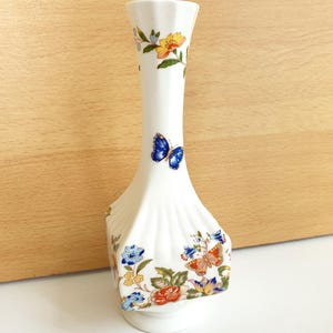 Puede incluir: Un jarrón de cerámica blanca con la parte superior acampanada y un diseño único con estrías. El jarrón está adornado con coloridos motivos florales y de mariposas en azul, amarillo y naranja. El jarrón mide unos 20 cm de alto.