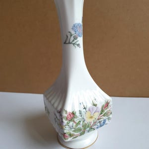 Puede incluir: Jarrón de porcelana blanca con borde y base dorados. El jarrón presenta un diseño floral con flores azules, rosas y amarillas. El jarrón tiene una forma geométrica única con una parte superior acampanada y una base cuadrada.