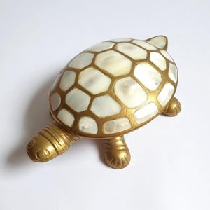 May include: A turtle-shaped decorative object featuring a gold-toned metal head, legs, and tail. The shell is composed of inlaid white, iridescent mother-of-pearl segments separated by a gold-colored metal grid pattern, creating a geometric design.