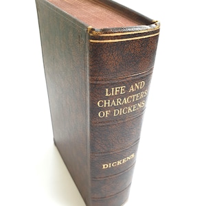Może przedstawiać: Brązowa książka w skórzanej oprawie z pozłacanymi napisami na grzbiecie. Tytuł to "Life and Characters of Dickens" z napisem "Dickens" umieszczonym pod tytułem.