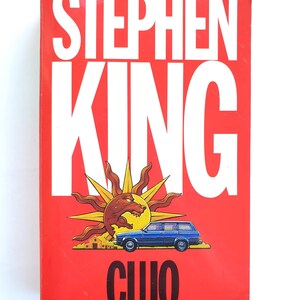 May include: A red book cover with the title "CUJO" by Stephen King. The cover features large white text, a sun graphic with a dog, and a blue station wagon.