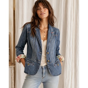May include: A blue denim blazer featuring vertical stitching, two brass-toned buttons, and two front flap pockets. The sleeves are rolled up to reveal a floral pattern lining in shades of red, green, and yellow. A white top is worn underneath.