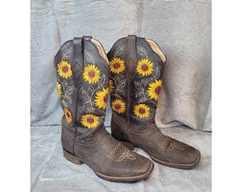 May include: A pair of dark brown leather cowboy boots featuring embroidered yellow sunflowers with green stems and leaves on the shaft. The boots have a square toe design and decorative stitching on the vamp, showcasing a classic western silhouette.