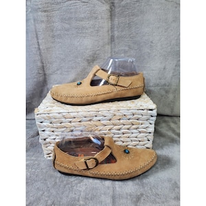 May include: A pair of tan leather shoes featuring a T-strap design with an adjustable brass-toned buckle, whipstitch detailing along the edges, and a small circular turquoise-colored inlay surrounded by radiating lines on the upper.