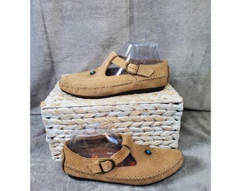 May include: A pair of tan leather shoes featuring a T-strap design with an adjustable brass-toned buckle, whipstitch detailing along the edges, and a small circular turquoise-colored inlay surrounded by radiating lines on the upper.
