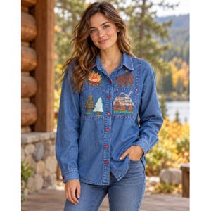 May include: A blue denim button-down shirt featuring embroidered patches of a campfire, a brown bear, two pine trees, and a log cabin. The shirt has a classic collar, long sleeves, and red buttons running down the front placket.