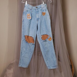 May include: Light blue denim jeans featuring a high-waisted fit with pleats at the waist. The jeans include three embroidered teddy bear patches and two large distressed holes at the knees, revealing the fabric underneath the denim material.