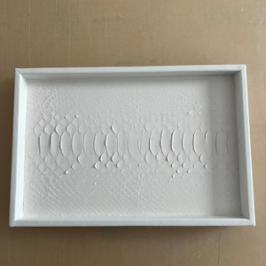 Könnte beinhalten: Ein rechteckiges, weißes Tablett mit einer strukturierten Oberfläche, die Schlangenschuppen ähnelt. Das Tablett hat einen erhöhten Rand und ist vor einem neutralen Hintergrund platziert. Die Textur ist ein herausragendes Merkmal.