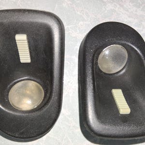 May include: Two black plastic car interior light fixtures. Each has a light-colored rectangular switch and a clear, round lens. The fixtures are oval-shaped with a slightly textured surface.