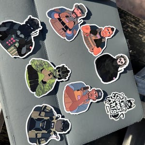 May include: A collection of colorful stickers featuring illustrations of military figures and a black cat. The stickers are on a gray tablet with a white stylus. The military figures are wearing tactical gear and holding weapons.
