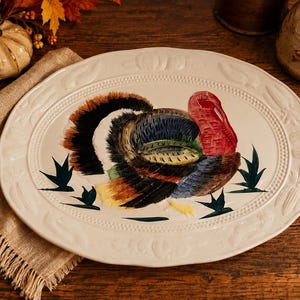 Puede incluir: Una fuente de servir ovalada de cerámica color crema presenta un pavo pintado a mano con plumaje vibrante en tonos marrón, negro, azul y rojo. El borde está grabado con un patrón de calabazas y hojas, realzado por un detalle de cuentas.