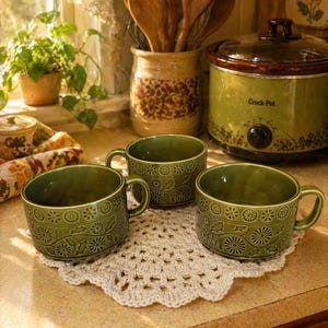 May include: Three green ceramic mugs with floral patterns sit on a cream-colored crocheted doily. A green Crock-Pot and a jar of wooden spoons are in the background. The scene is set in a kitchen with natural light.