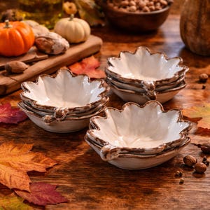 May include: A stack of white leaf-shaped ceramic bowls with a brown rim and handle. The bowls are on a wooden table with pumpkins, nuts, and fall leaves. The bowls are perfect for serving snacks or dips.