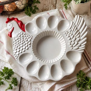 Puede incluir: Una bandeja de cerámica blanca para servir huevos con forma de gallina, con cresta y barbillas rojas. La bandeja presenta una sección central circular con borde estriado rodeada por doce hendiduras ovaladas para los huevos.